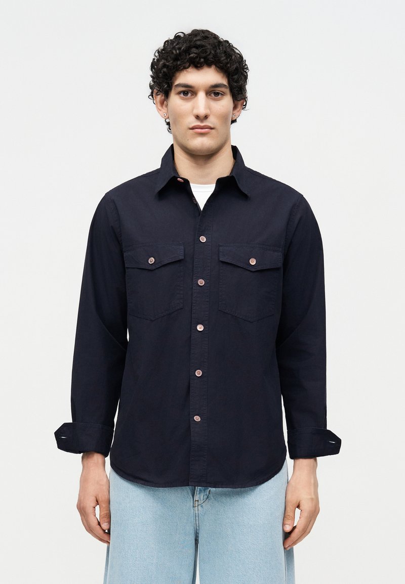 Άνδρας με σγουρά μαλλιά που φοράει σκούρο μπλε πουκάμισο με τσέπες στο στήθος και ανοιχτόχρωμα τζιν, στέκεται μπροστά σε ένα μονόχρωμο φόντο.