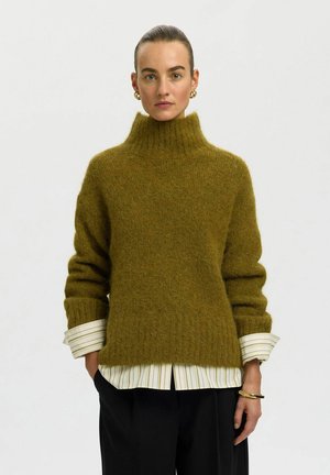 Mujer con suéter de cuello alto peludo color verde oliva sobre camisa a rayas y pantalones negros, con pendientes de aro dorados y pulsera, fondo neutro.