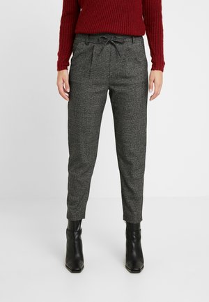 Kvinde iført grå ternede bukser med snørebånd i taljen, sorte ankelstøvler og en rød strikket sweater, stående mod en ensfarvet baggrund.