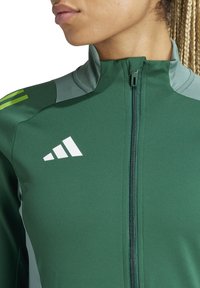 Grüne Sportjacke mit Reißverschluss vorne, hohem Kragen und kontrastierenden hellgrünen Schulterakzenten; beinhaltet ein Logo mit drei weißen Streifen.