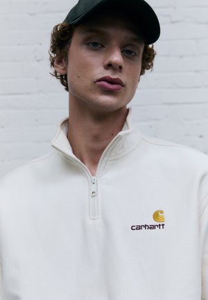 Pull en polaire blanc avec un col zippé à mi-hauteur, arborant un logo brodé jaune et bordeaux. Texture lisse et coupe moderne.