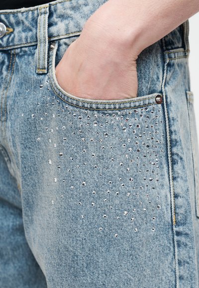 Χέρι σε μπροστινή τσέπη από ανοιχτό γαλάζιο τζιν διακοσμημένο με μικρές ασημένιες τρουκς.