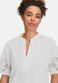 Blusa bianca in cotone con vestibilità comoda, caratterizzata da un profondo scollo a V e maniche corte, decorata con ricami floreali intricati lungo i polsini.