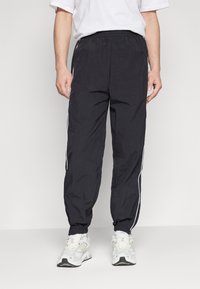 Zwarte sportieve broek van een lichtgewicht, gestructureerde stof, met witte zijstrepen en elastische boorden, gedragen met witte sneakers.