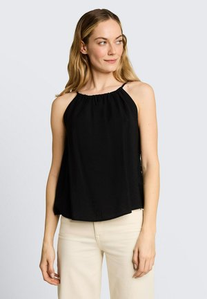 Femme aux longs cheveux blonds portant un haut noir sans manches à col licou et un pantalon beige clair, regardant vers sa droite sur un fond clair.