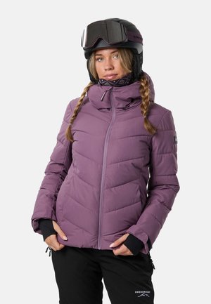 Une femme portant une veste de ski violette isolante, un pantalon noir et un casque avec des lunettes de ski pose devant un fond blanc uni.