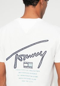 Λευκό βαμβακερό T-shirt με γυαλιστερό λογότυπο "Tommy" σε ναυτικό μπλε και σχέδιο κειμένου σε τυρκουάζ με ονόματα πόλεων στην πλάτη.