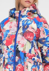 Giacca imbottita con motivo floreale in vivaci colori rosso, blu e verde. Presenta una cerniera blu e tasche per la funzionalità.