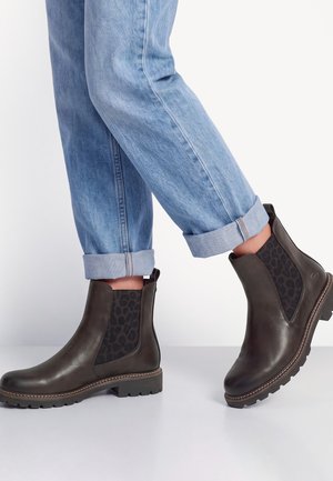 Personne portant un jean bleu clair roulé et des bottines en cuir marron foncé avec des panneaux latéraux élastiques à imprimé léopard.