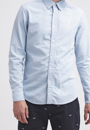 Hellblaues, langärmliges Hemd mit Knöpfen, getragen zu schwarzen Hosen mit kleinen, weißen Stickereien.