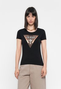 Kvinde med strit sort hår bærer sort Guess T-shirt med metallisk trekantlogo og beige vide bukser foran en hvid baggrund.