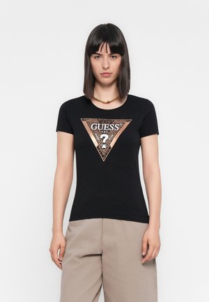 Mujer con cabello lacio negro que lleva una camiseta negra de Guess con un logo metálico en forma de triángulo y pantalones de pierna ancha beige, frente a un fondo blanco.