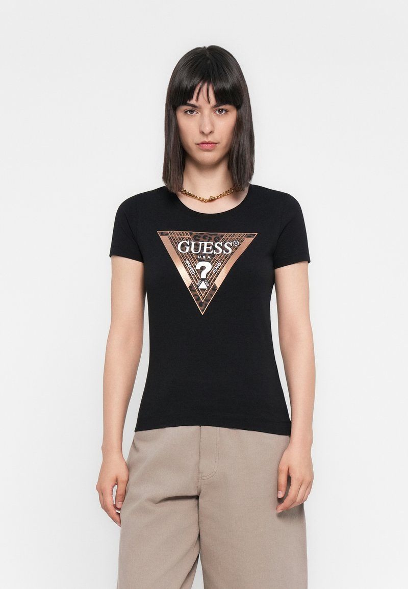 Vrouw met stijlvol zwart haar draagt een zwarte Guess t-shirt met een metalen driehoeklogo en beige wijde broek op een witte achtergrond.