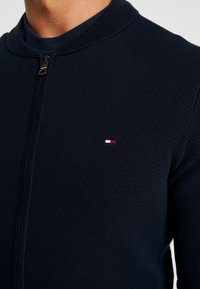 Námořnická svetr s czipem, vyrobený z texturované pleteniny, s vysokým límcem a malým vícetrojbarevným logem na prsou.