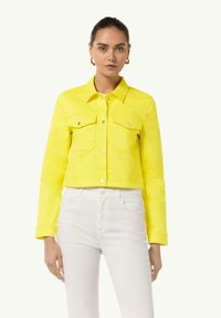 Yellow cropped denim jacket with a button-up front, two chest pockets, and a classic collar; paired with white jeans.