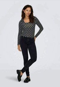 Γυναίκα που στέκεται με το ένα χέρι στη μέση, φορώντας μαύρα τζιν, μαύρο πουκάμισο με λευκές βούλες και μαύρες λοφάδες σε απλό φόντο.