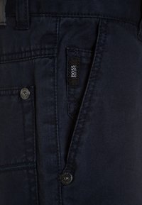 Námořnické modré kalhoty vyrobené z husté látky, s viditelnou kapsou a černou nálepkou s nápisem "BOSS" a detaily ve stříbrné barvě v podobě patentního knoflíku.