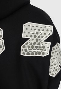 Schwarzer Stoff mit drei großen grauen und weißen Patchbuchstaben, die hexagonale und Punktmuster aufweisen und aufgenäht sind.