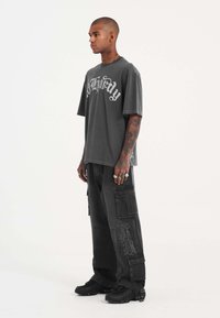Camiseta gris de gran tamaño con texto gráfico blanco, combinada con pantalones cargo negros que tienen bolsillos y detalles bordados. Zapatillas negras.