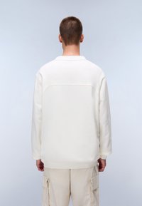 White, long-sleeve sweatshirt with a crew neck, smooth texture, and a seam detail across the upper back. Paired with beige cargo pants.