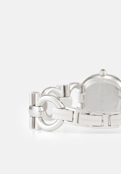 Silver metallarmband klocka med rund boett, länkdesign och graverade logotypdetaljer på remmen. Slät, polerad yta.