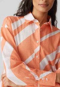 Camicia a righe arancioni e bianche, con colletto, maniche lunghe e una texture del tessuto che assomiglia al lino. Presenta piccoli bottoni trasparenti.