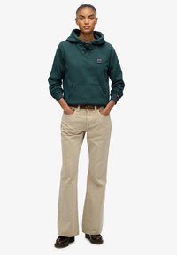 Sudadera con capucha de color verde oscuro y bolsillo frontal, combinada con pantalones acampanados beiges. El conjunto se complementa con zapatos oscuros y un cinturón de estampado.