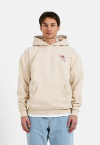 Beige hoodie med en framficka, dragsko kapuschong och en broderad logotyp i röd. Avslappnad passform med ribbad mudd och nederkant. Buren över en vit skjorta.