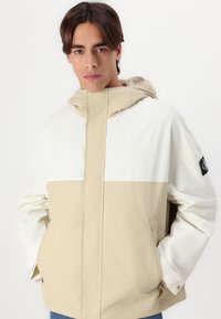 Jachetă de vânt bej și albă, cu glugă, închidere frontală cu fermoar și buzunare laterale. Material texturat cu design în blocuri de culoare.