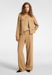 Beige skräddarsydd kostym med en kort jacka med dubbelbröstfickor och matchande vida byxor. Mjuk textur, minimala hårdvarudetaljer.