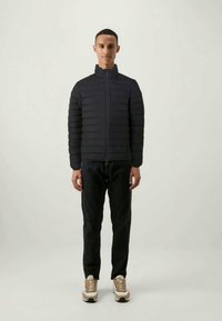 Sort puffer-jakke med horisontalt quiltet design, høj krave, fuld frontlynlås; parret med mørke bukser og lyse sneakers.