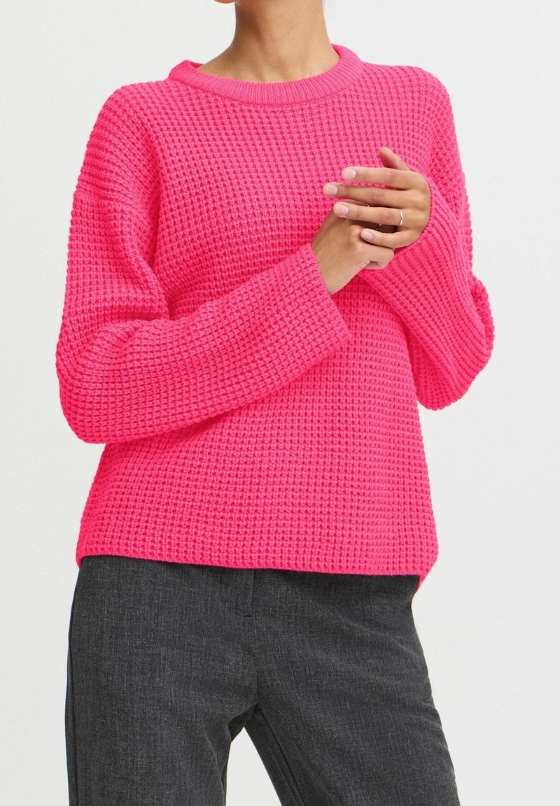 Pull en tricot rose avec un motif texturé, col rond et manches longues, associé à un pantalon gris foncé. Le pull a une coupe décontractée.