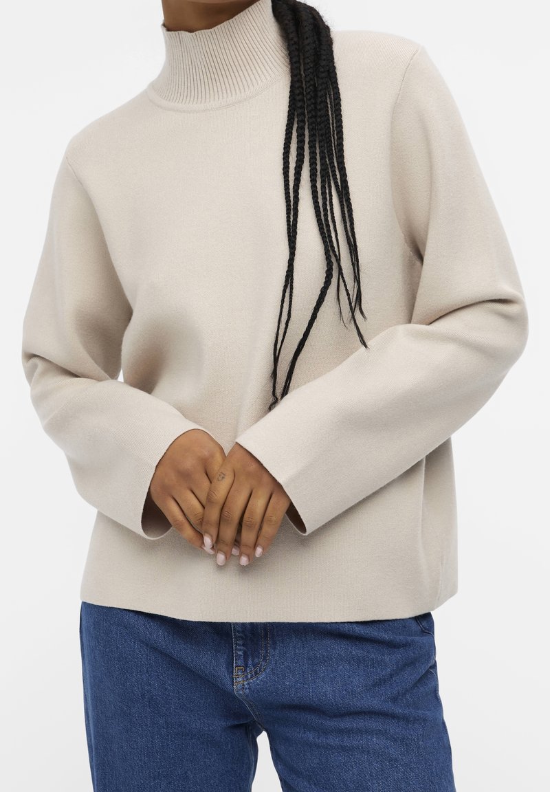 Beige, geribbelde coltrui met een rechte snit en lange mouwen, gecombineerd met blauwe denim jeans. De textuur lijkt zacht en glad.