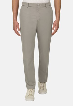 Homme portant un pantalon gris clair à jambes droites et des baskets beiges, debout devant un fond uni.