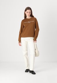 Hnědá mikina s bílým logem "TOMMY HILFIGER", v kombinaci s bílými kalhotami a černými botami. Lehká látka, kulatý výstřih, volný střih.