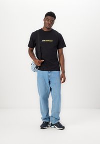 Black cotton T-shirt with yellow and white logo, light blue denim pants with large pockets, and black sneakers with blue accents.