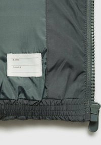 Sezione di giacca imbottita grigia con un'etichetta in tessuto bianco e righe per nome e telefono. Include orlo elasticizzato e zip laterale.