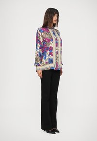 Kvinde står i sorte bukser og en langærmet skjorte med knapper, der har dristige blomster- og leopardtryk i lilla, blå og beige nuancer.
