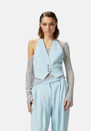 Chaleco corto azul claro con un escote profundo, tres botones delanteros y dos bolsillos laterales, combinado con una blusa plateada de manga larga y pantalones de pierna ancha.