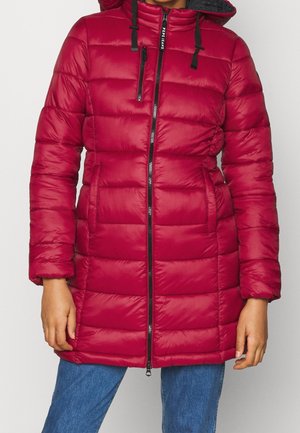 Mujer con una chaqueta acolchada roja con capucha, cremallera delantera y bolsillos, combinada con jeans azules frente a un fondo simple.