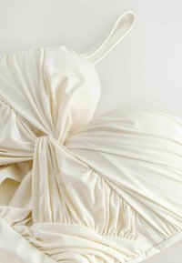 Haut de maillot blanc avec tissu froncé et détail noué au centre. Confectionné dans un matériau lisse avec des bretelles fines et une forme structurée.