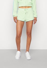 Žena v pastelově zelených kraťasech na šňůrku a odpovídajícím zkráceném sáčku se bílými teniskami, stojící před jednoduchým bílým pozadím.