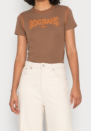Bruine T-shirt met oranje "BDG JEANS" tekst, aansluitend ontwerp, korte mouwen en contrasterende stiksels. Gecombineerd met hooggetailleerde, roomkleurige jeans.