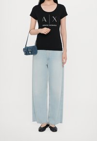 Camiseta negra de manga corta con logo plateado, jeans de pierna ancha azul claro y un pequeño bolso de mezclilla azul. Bailarinas negras. Atuendo simple y casual.
