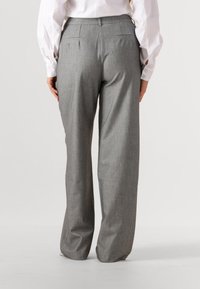 Vrouw die grijze wijde pantalons met krijtstreep en een wit shirt met lange mouwen draagt, gezien van achteren tegen een effen achtergrond.