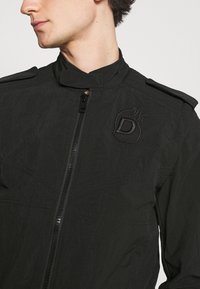 Černá zipová bunda s vysokým límcem, strukturovanou tkaninou a decentním logem. Obsahuje akcenty na ramenou a zúžený siluetu.