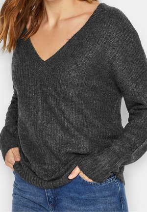 Femme portant un pull en tricot côtelé gris foncé à col en V avec manches longues et jean bleu, les mains dans les poches.