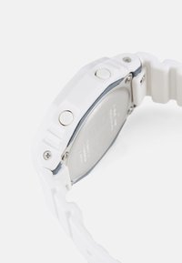Montre numérique en caoutchouc blanc avec un cadran rectangulaire, deux boutons latéraux et une finition mate. Le dos est en métal argenté avec du texte gravé.