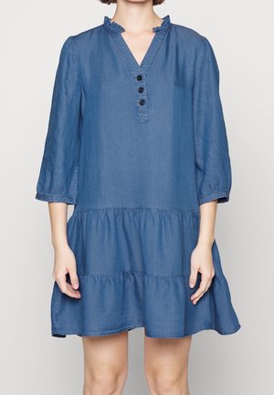 Kobieta ubrana w luźną niebieską sukienkę dżinsową z rękawami do łokci, z falbankowym kołnierzykiem, trzema czarnymi guzikami i wielowarstwową spódnicą.