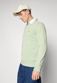 Man in een lichtgroene sweatshirt boven een wit overhemd, met een crème pet en blauwe spijkerbroek, staande tegen een effen lichte achtergrond en naar beneden kijkend.